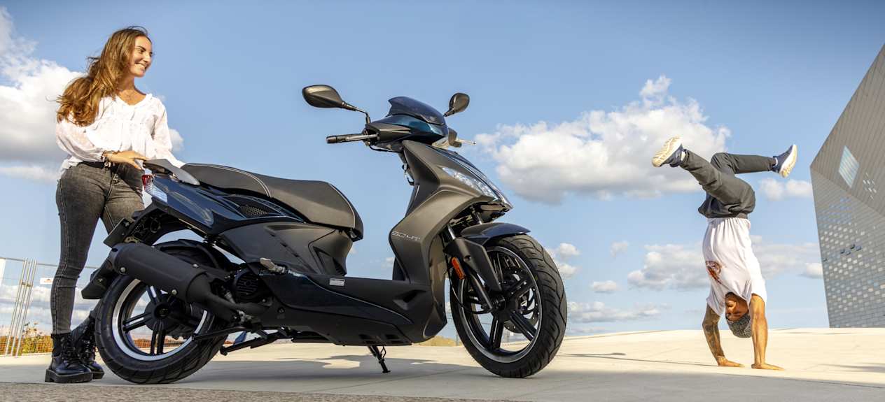 Kymco Agility 50 16+ 2024 : profil droit statique avec jeune fille et figure de hip hop