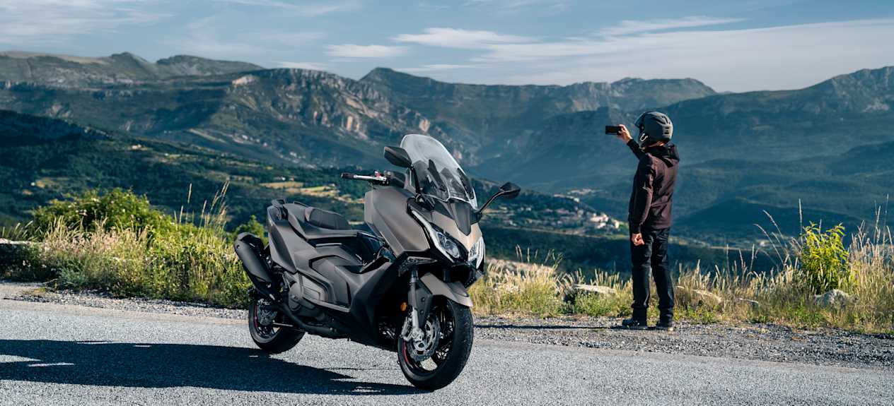 Kymco AK Premium 2024 : trois quarts avant droit avec vue sur montagnes et motard