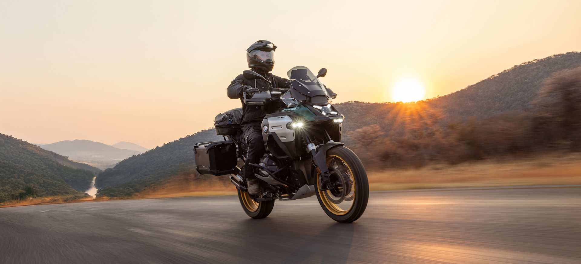 A motorcyclist rides on a winding road at sunset, surrounded by hills and a river.