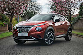 A red Nissan Juke parked on a tree-lined street with blooming flowers in the background.