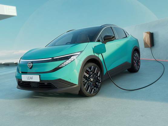 A turquoise electric Nissan LEAF parked near a charging station by the sea, with a rocky backdrop