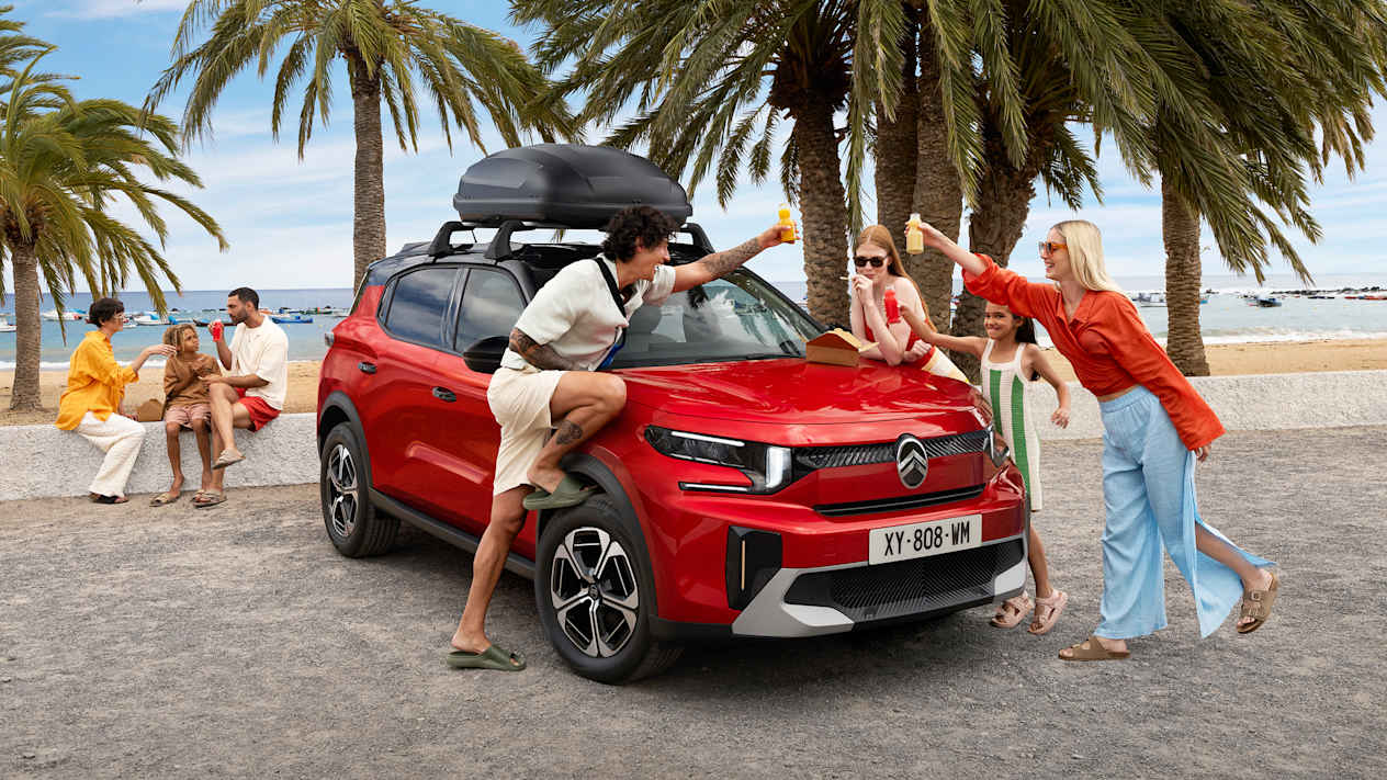 Citroën C3 Aircross 2024 : trois quarts avant droit statique en bord de plage avec coffre de toit et famille