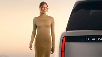 A woman in a beige dress stands beside a luxury Range Rover Electric vehicle against a soft sunset 