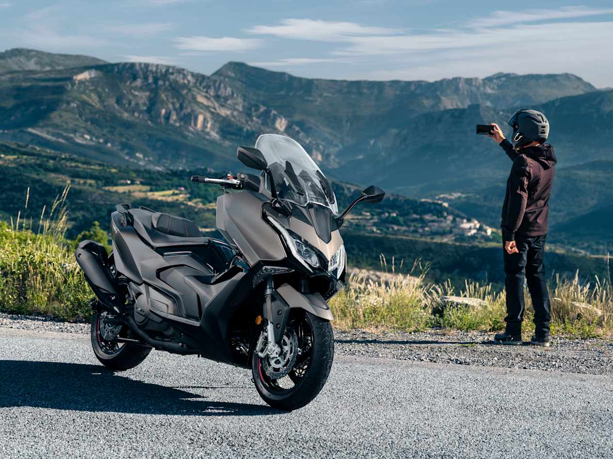 Kymco AK Premium 2024 : trois quarts avant droit avec vue sur montagnes et motard