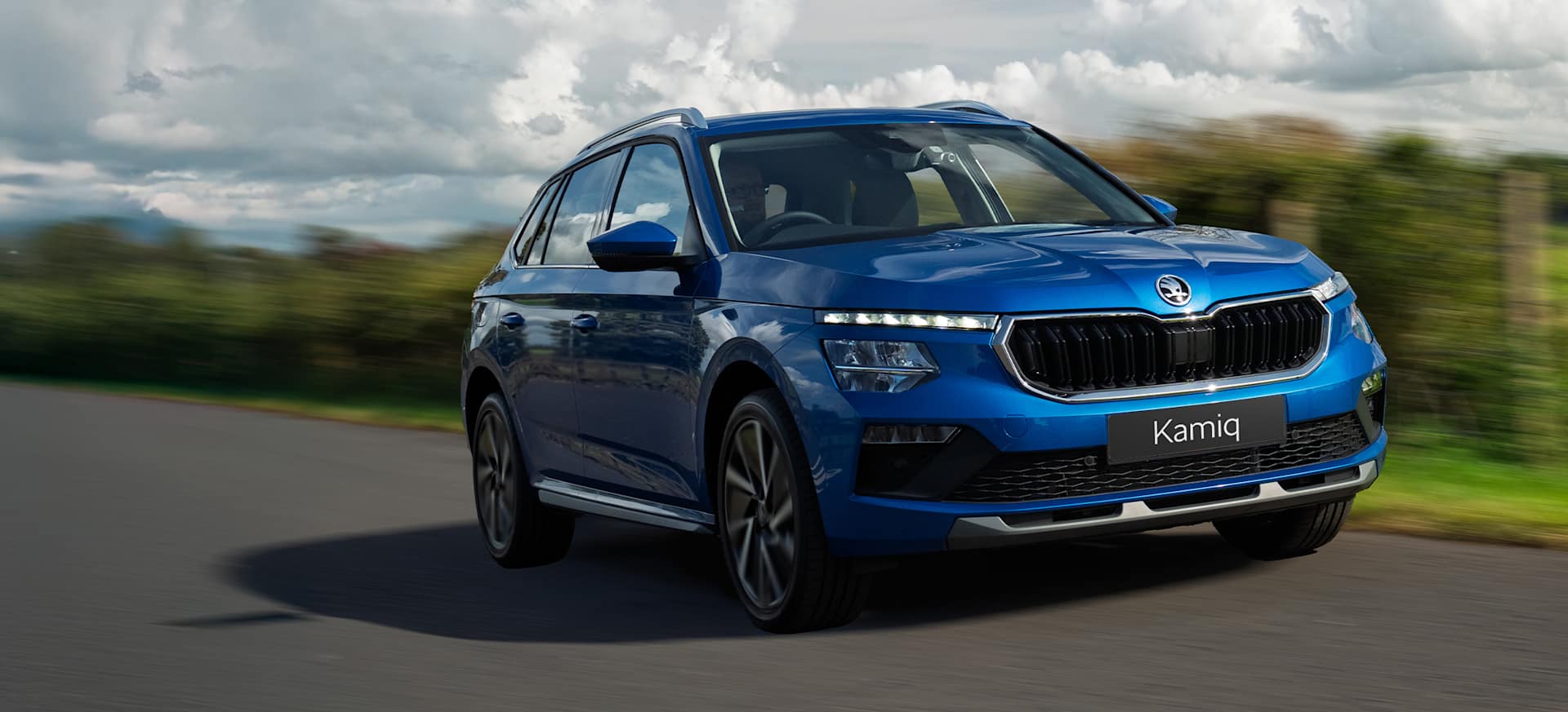 A blue Škoda Kamiq driving on a winding road surrounded by greenery under a cloudy sky.