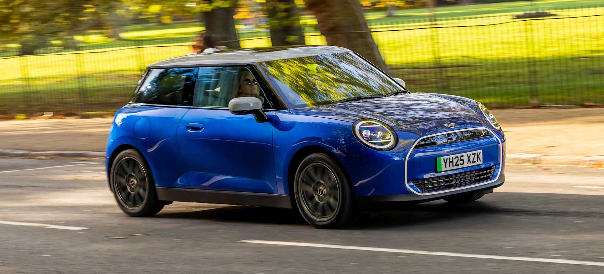A blue MINI car driving on a road with trees in the background.