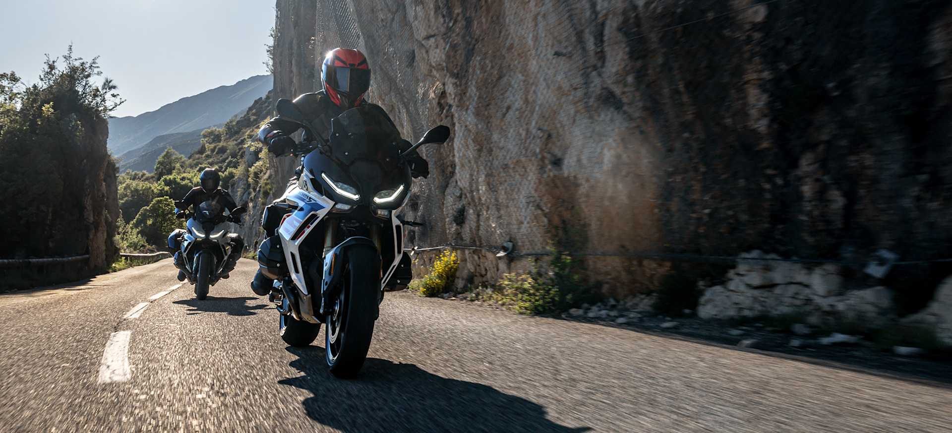 Two motorcyclists riding on a winding road surrounded by rocky cliffs and greenery.