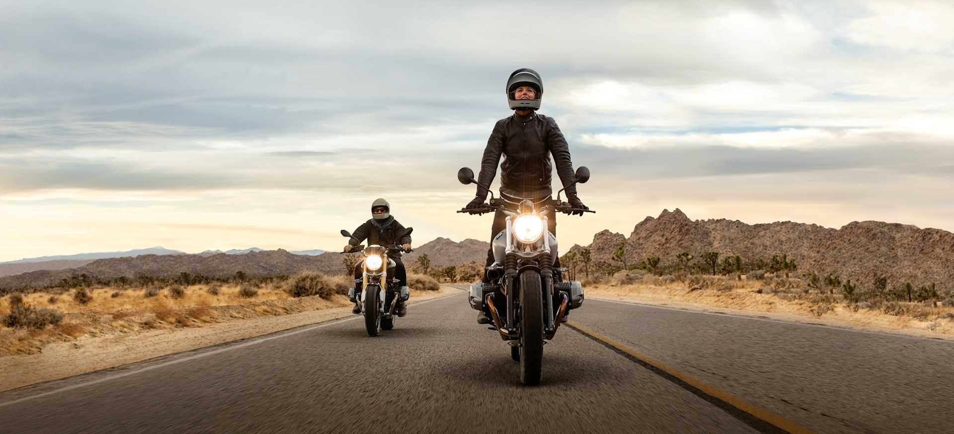 Two motorcyclists riding on a winding road through a desert landscape under a cloudy sky.