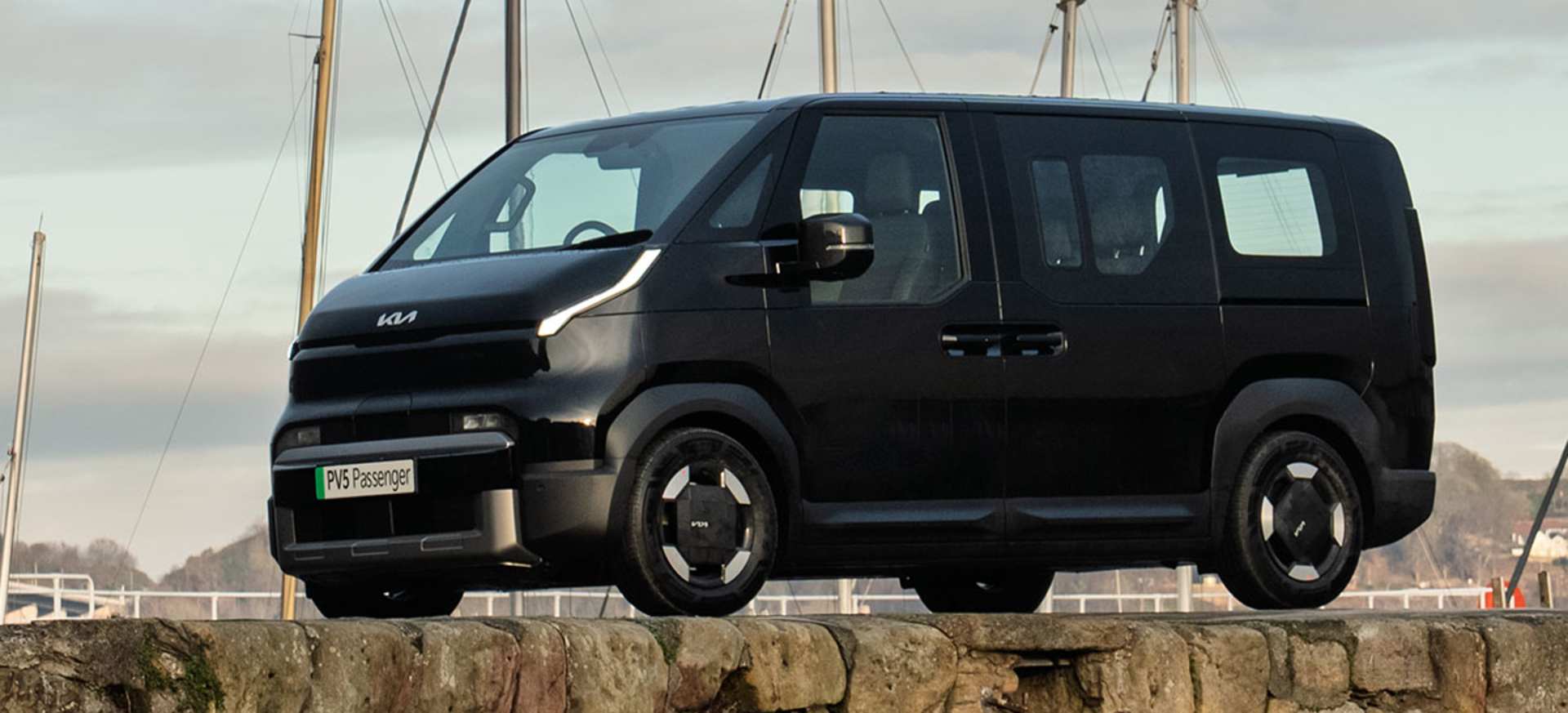 A black Kia PBV 5 Passenger parked on a stone wall with boats in the background.
