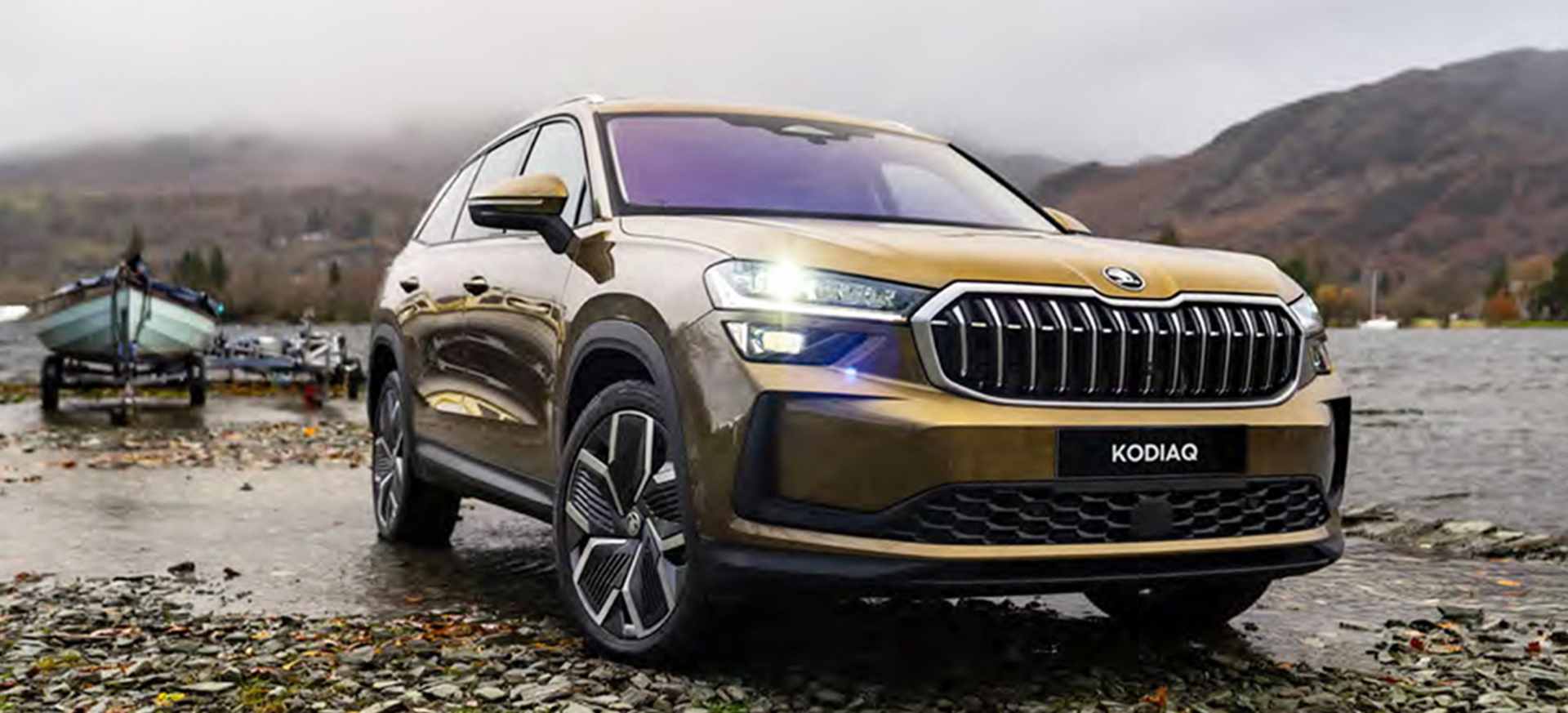A Škoda Kodiaq SUV parked by a lake with boats in the background and mountains in the distance