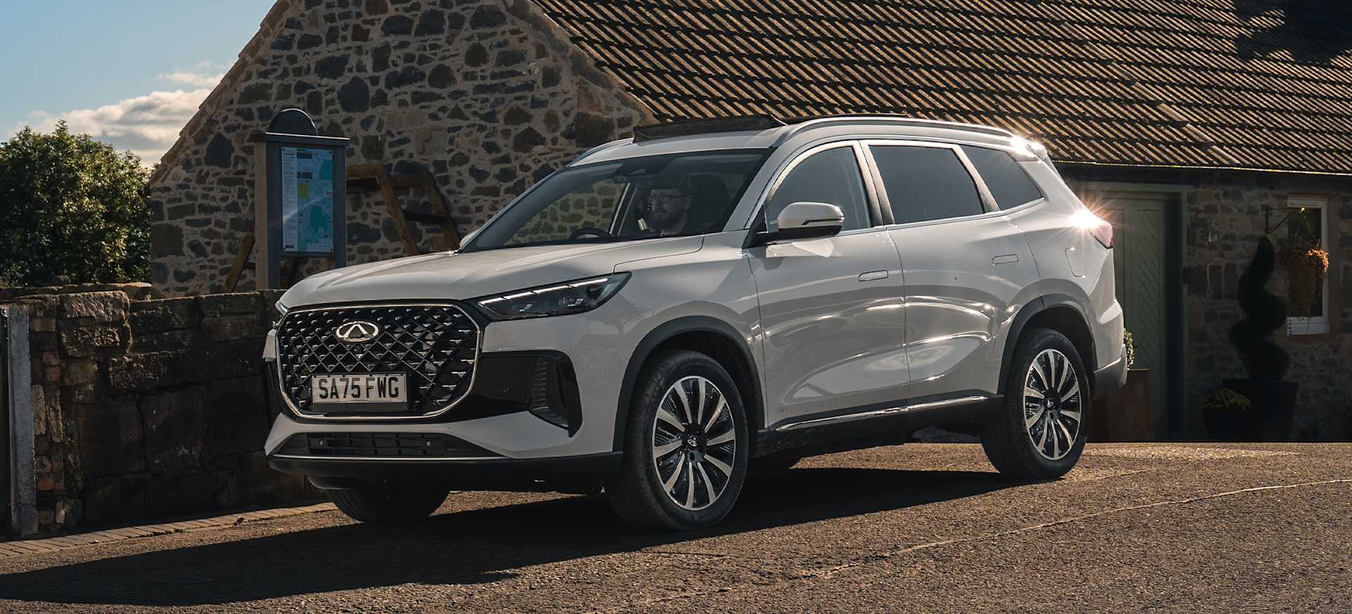A modern white Chery TIGGO 8 parked near a stone cottage with greenery in the background.
