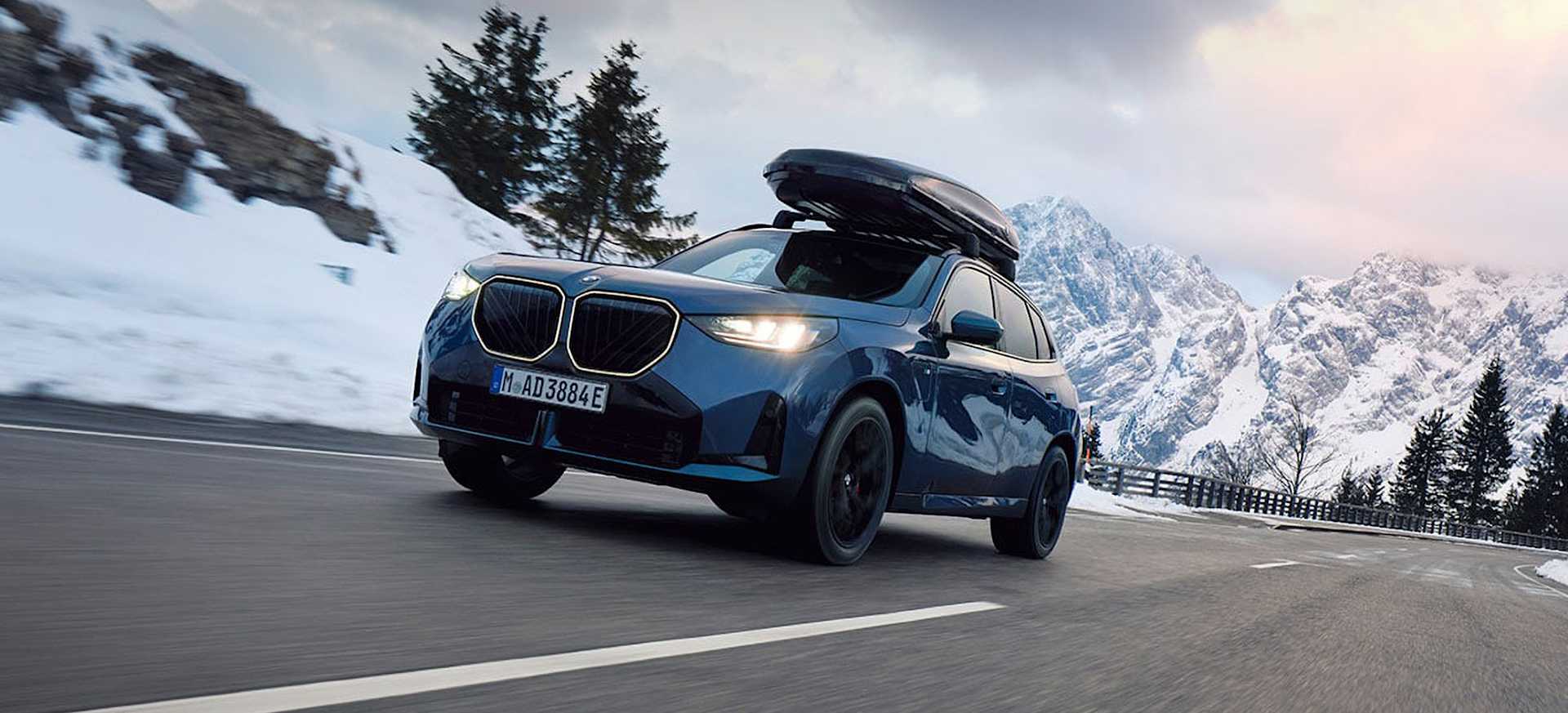 A blue BMW with a roof box drives along a snowy road with mountains in the background.