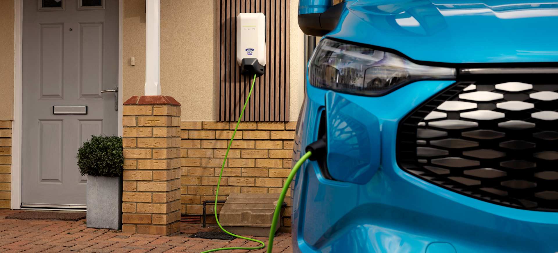 A blue electric Ford Transit Van charging at a home with a wall-mounted charger and a door in the background.
