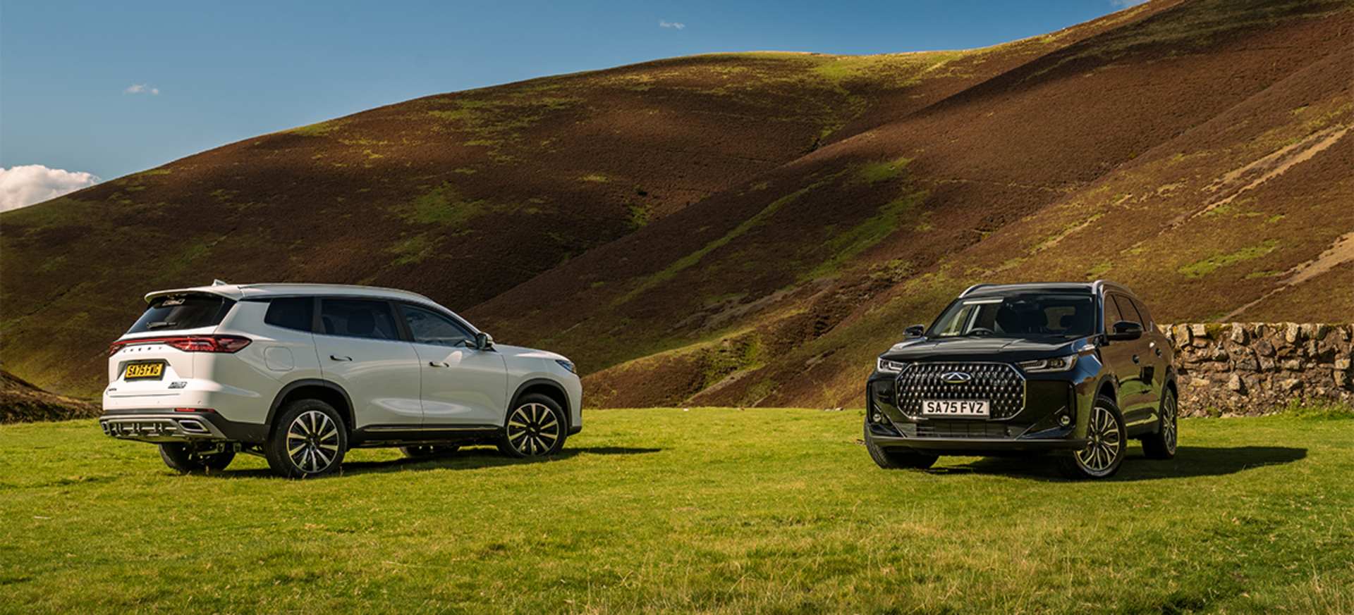 A black Chery TIGGO 7 & a white Chery TIGGO 8 parked on green grass with a hillside backdrop.