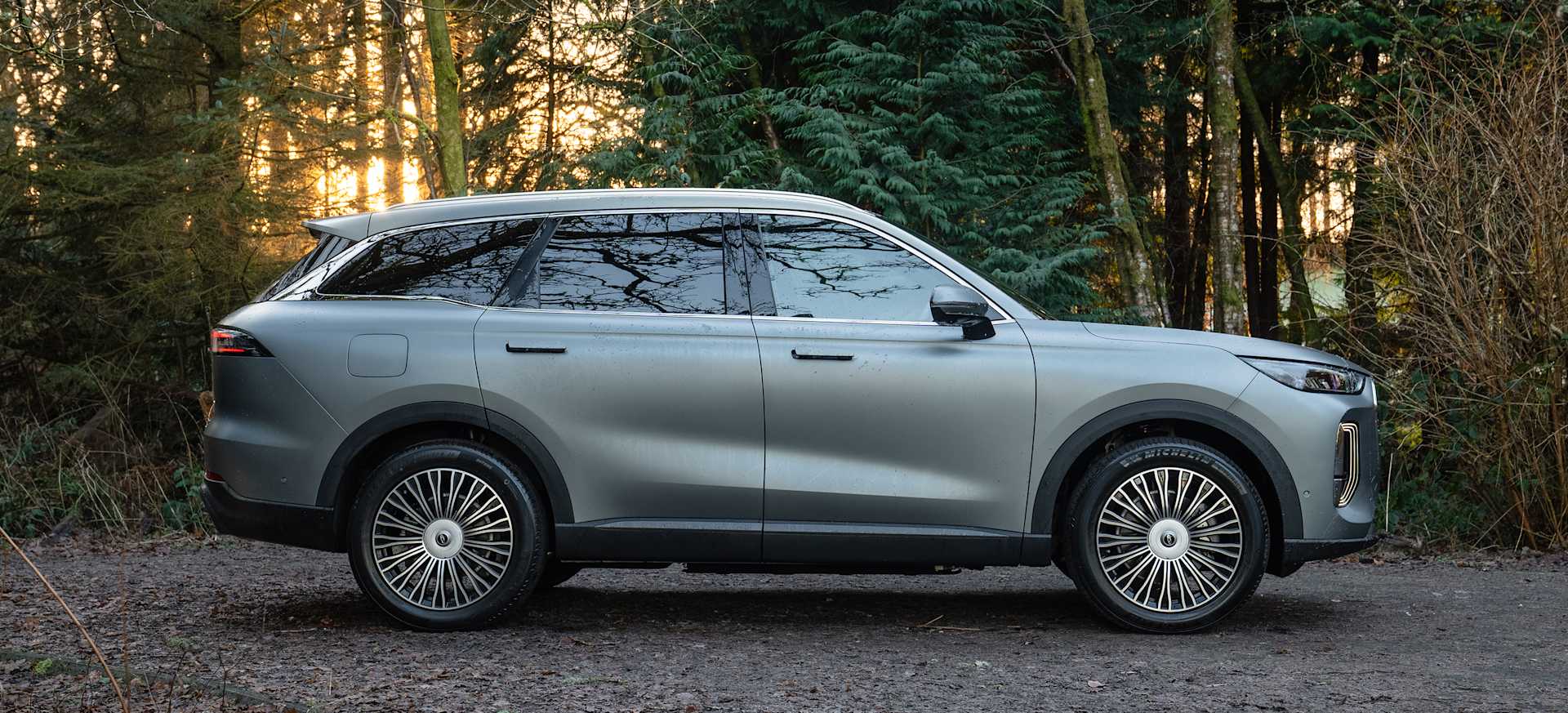 A sleek Chery TIGGO 9 parked on a gravel path surrounded by trees at sunset.