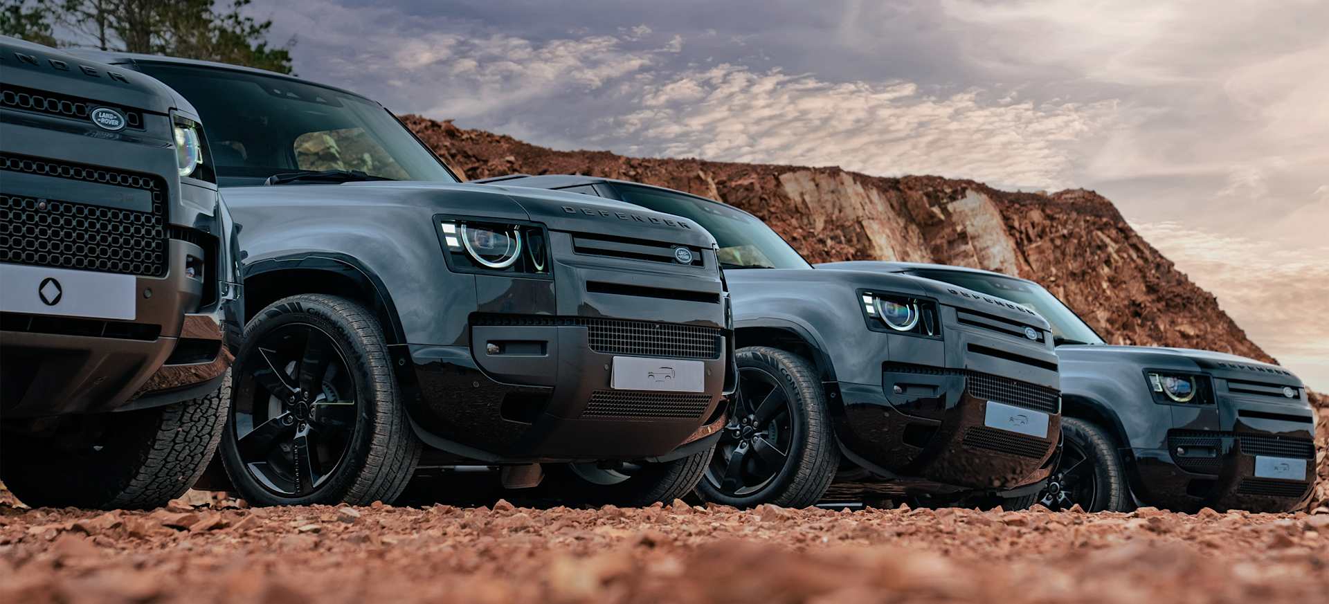 A lineup of rugged Defender SUVs parked on rocky terrain under a dramatic sky.