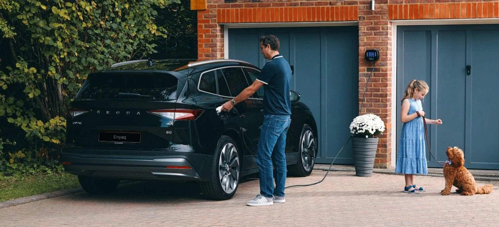 A man charges a black Škoda Enyaq while a girl and a dog stand nearby in a driveway.