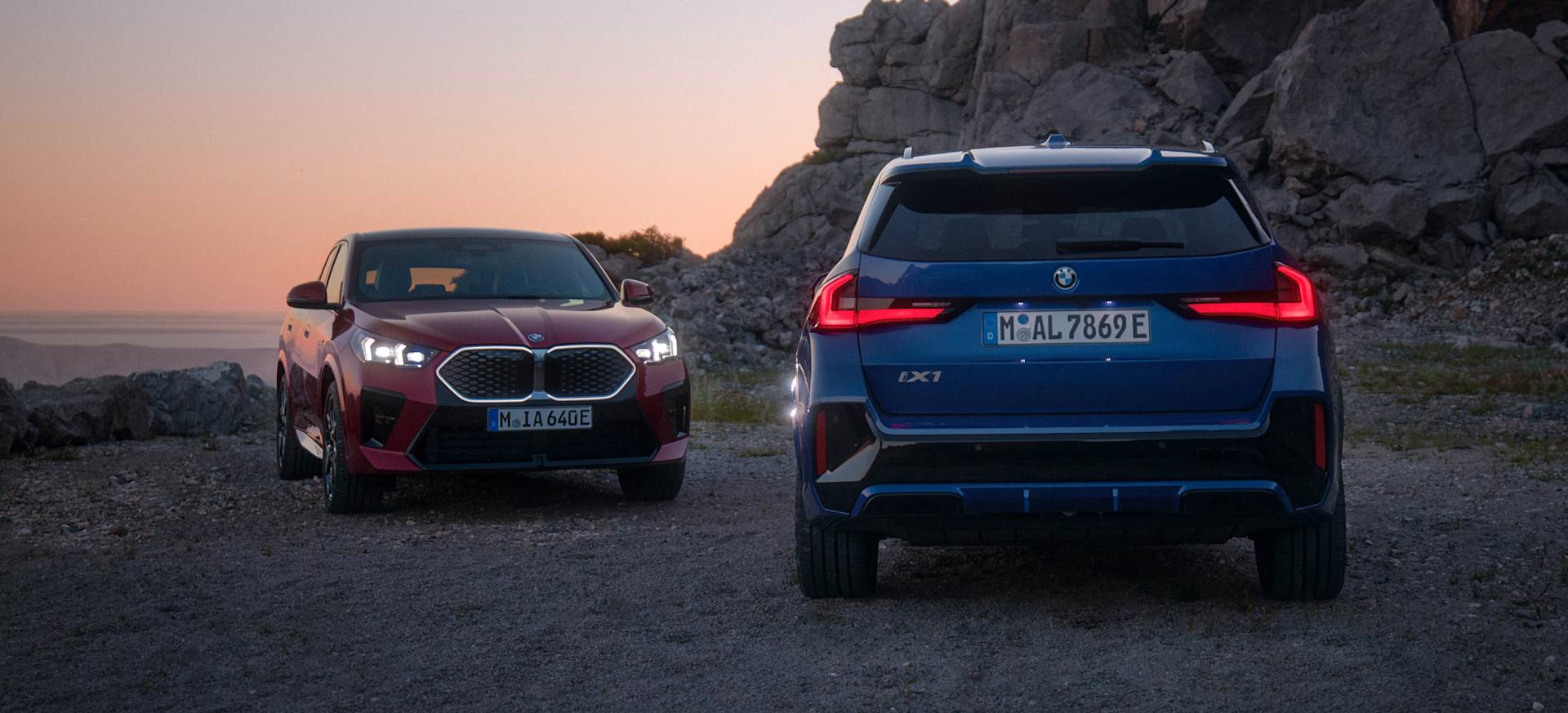Two BMW cars, one red and one blue, parked on rocky terrain at sunset.