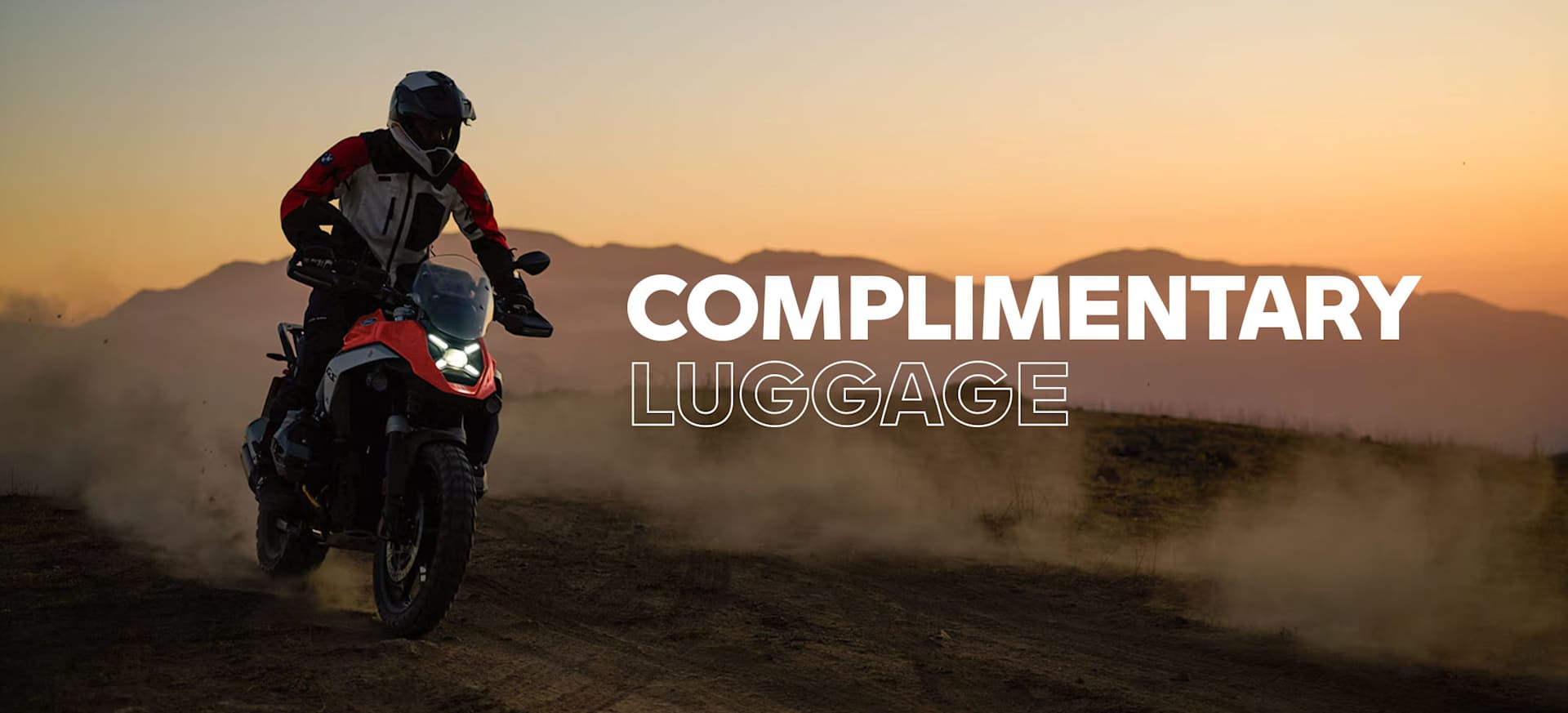 A motorcyclist riding on a dirt path at sunset, with the text "COMPLIMENTARY LUGGAGE" overlayed.