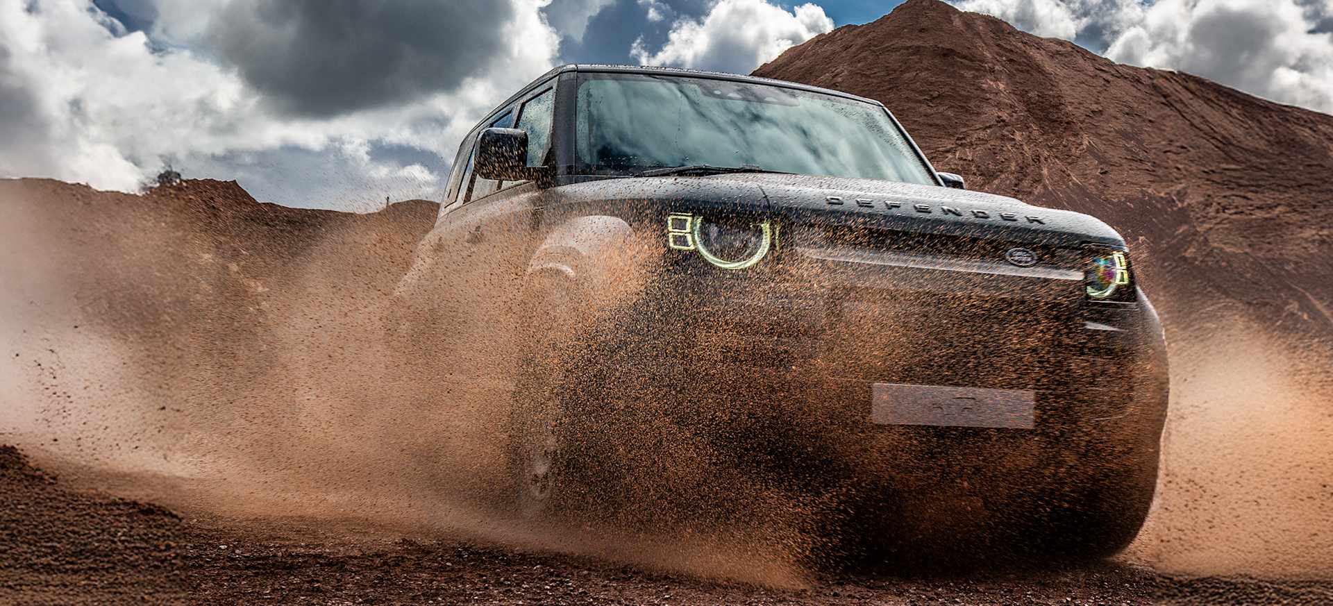 A rugged Defender OCTA kicking up dust while driving on a dirt track under a cloudy sky.