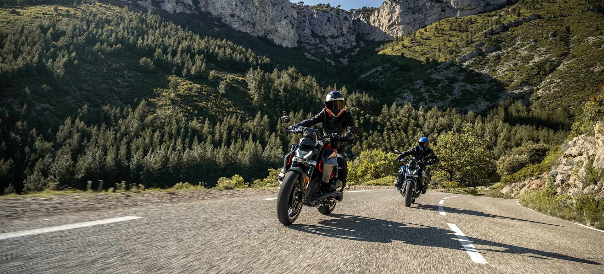 Two motorcyclists riding on a winding road surrounded by lush green hills and trees.