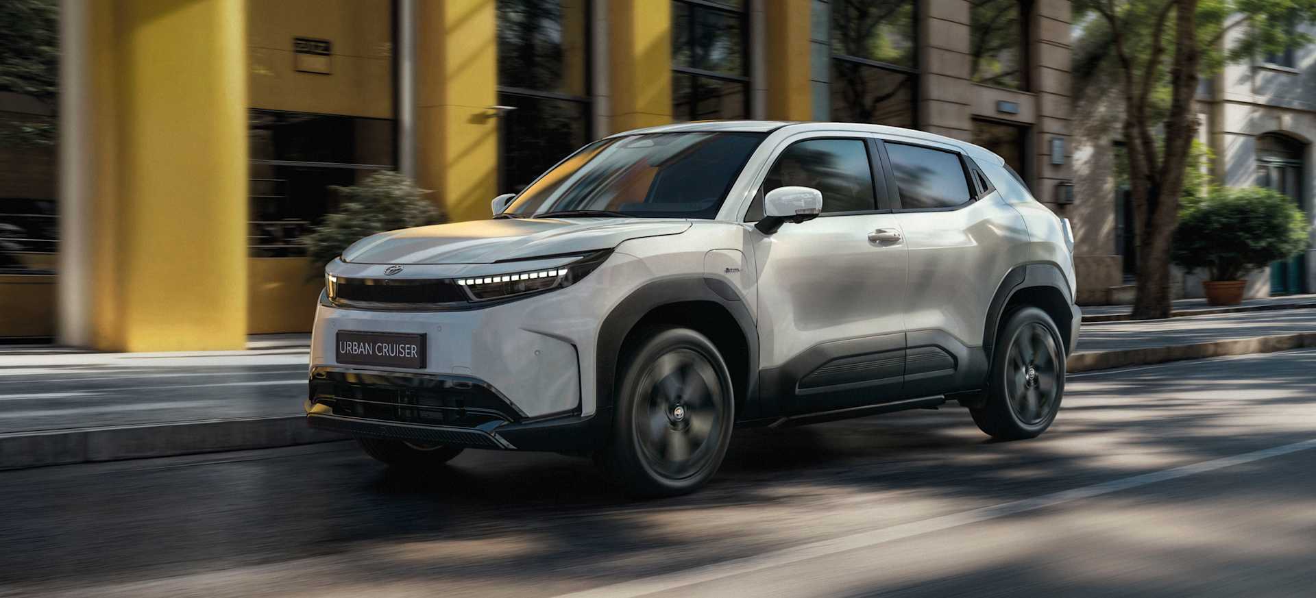 A modern white SUV driving along a city street with trees and buildings in the background.