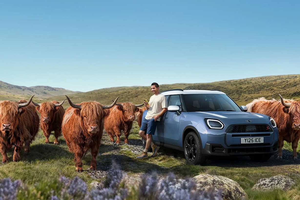 A man stands beside a blue car, surrounded by Highland cattle in a scenic landscape.