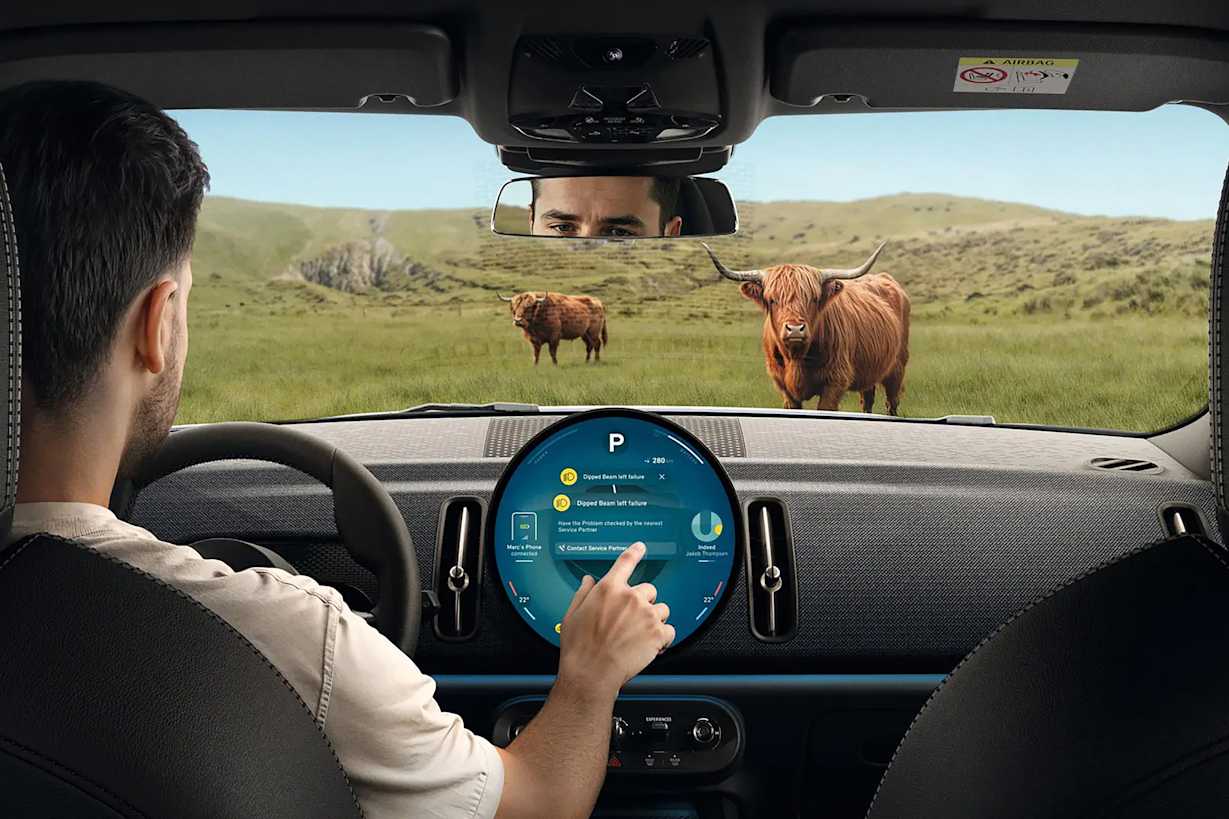 A person inside a car interacts with a digital display while Highland cattle graze outside.