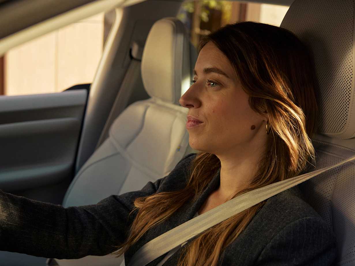 A woman with long hair sits in a car, looking ahead while wearing a seatbelt.