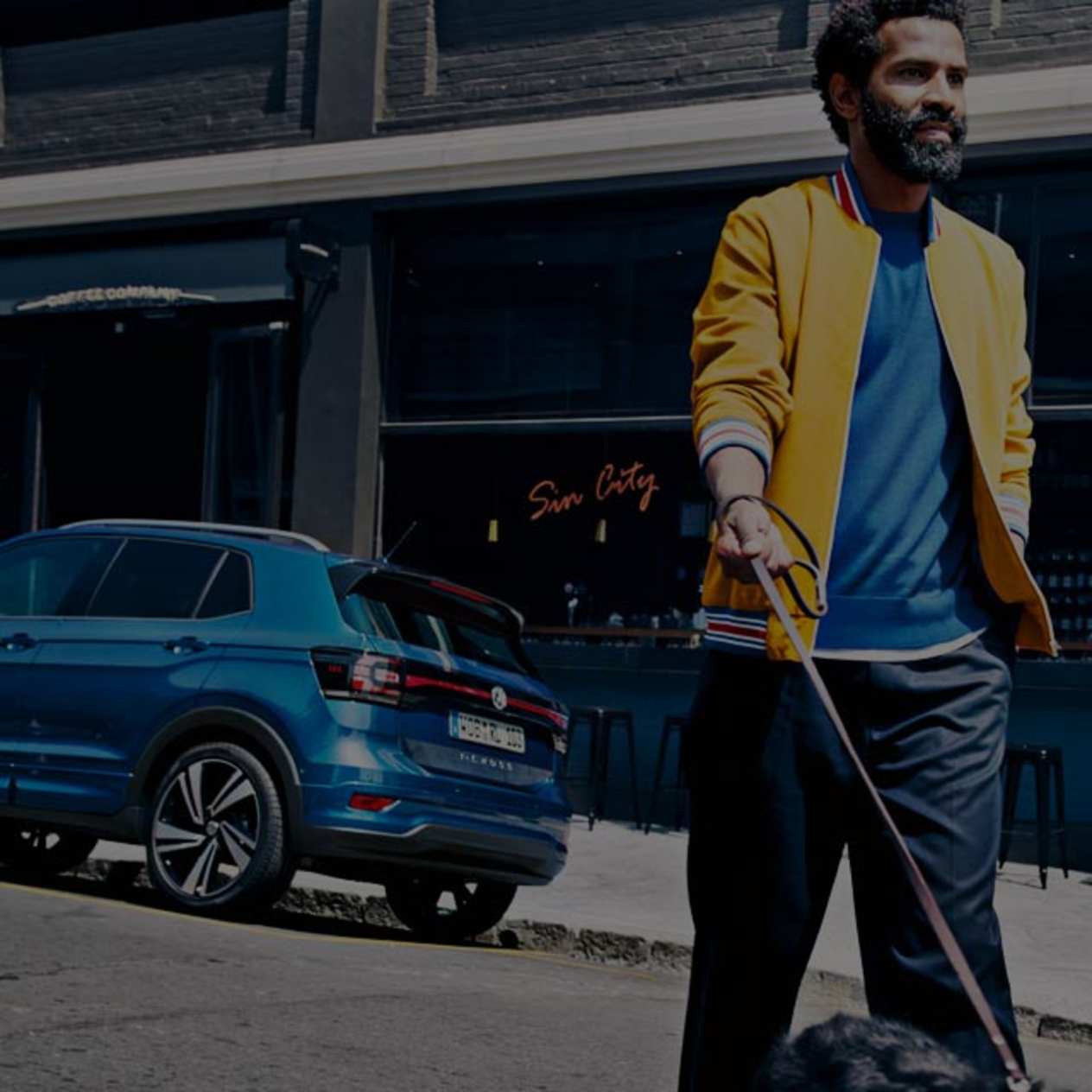 Man in yellow jacket walking dog in the opposite direction of a blue Volkswagen