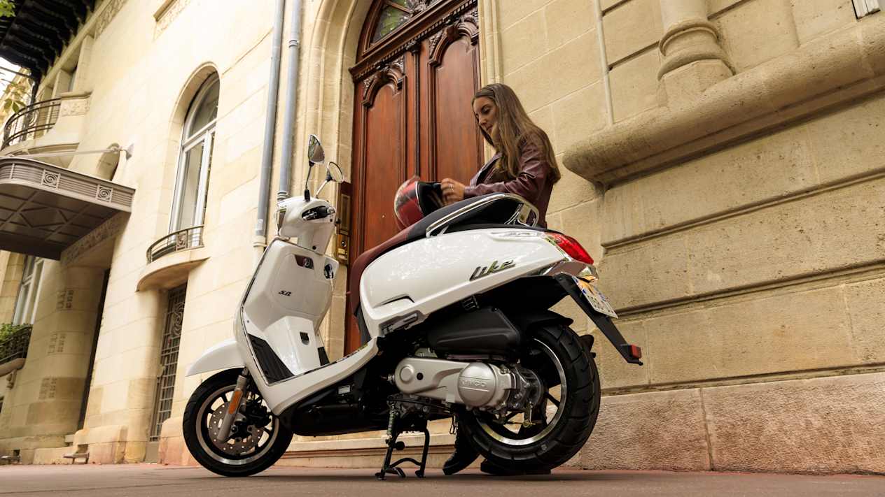 Kymco Like 50 2024 : trois quarts arrière gauche statique avec jeune femme et bâtiment ancien