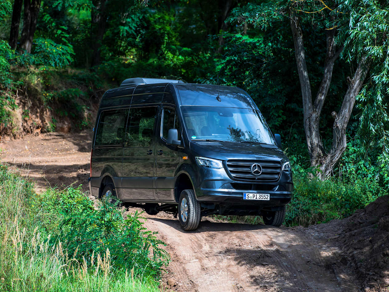 A dark grey Mercedes Sprinter drives along a muddy road showing the side and front of the van