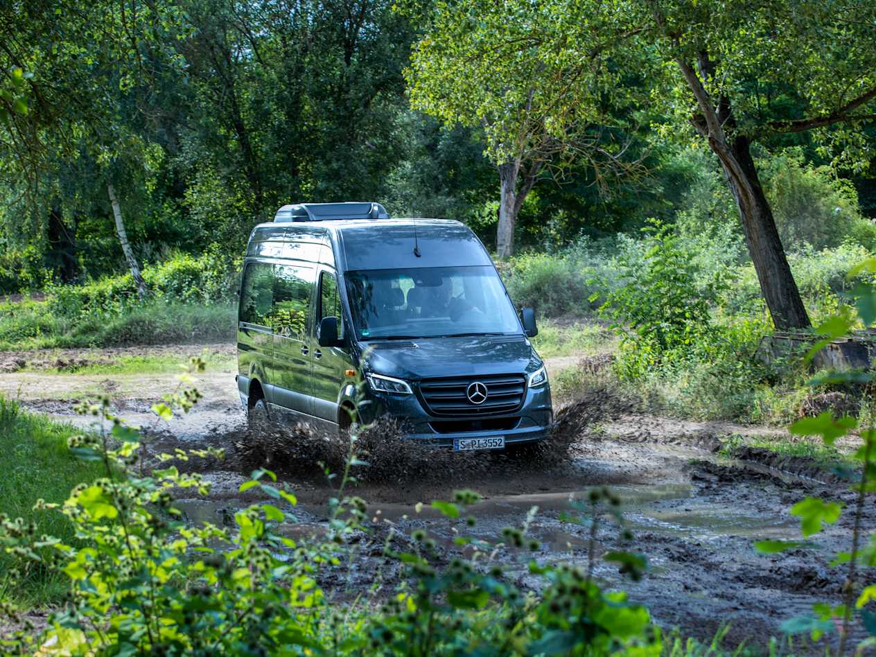 A dark grey Mercedes Sprinter drives through water in a wooded area