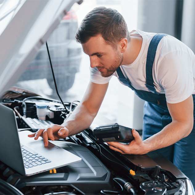 A car mechanic in white shirt and blue overalls using a laptop to run diagnostics while holding a tool in his hand.